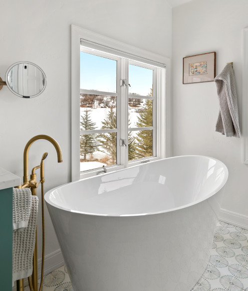 Modern bathroom with freestanding white tub, brass faucet, patterned tile floor, and snowy mountain view